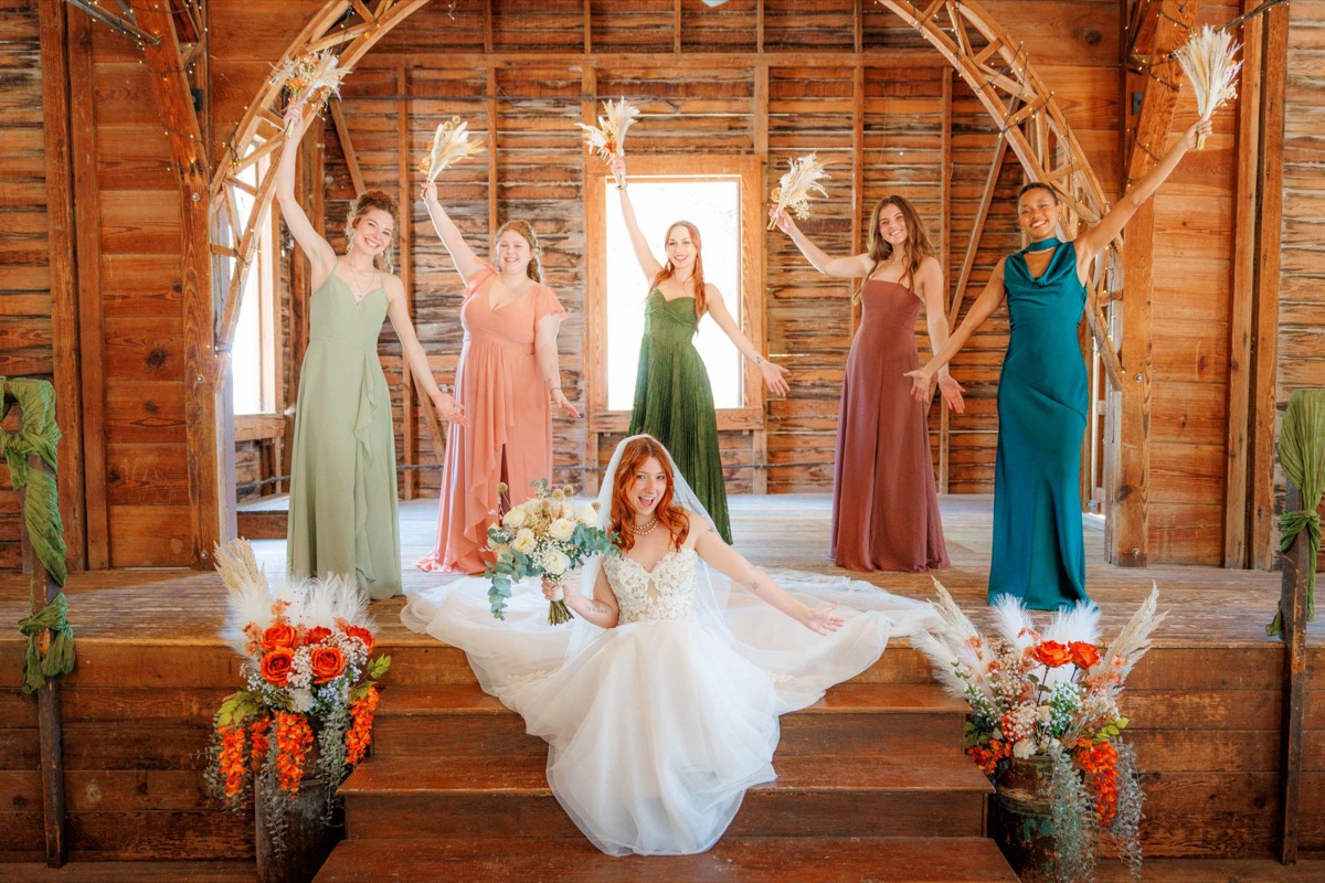 A bride sits on the barn stage steps as her bridesmaids raise their bouquets behind her