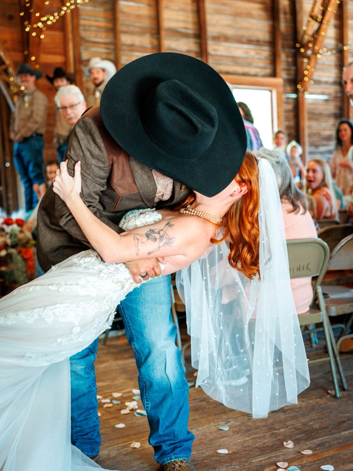 The groom in a black cowboy hat dips his bride into a kiss as guests cheer