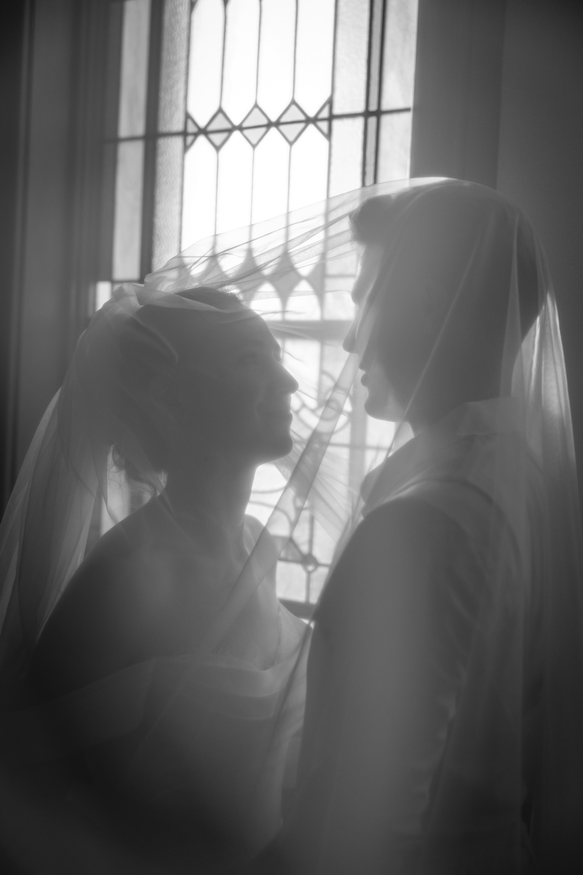 A bride and groom face each other beneath her veil by a window, in black and white
