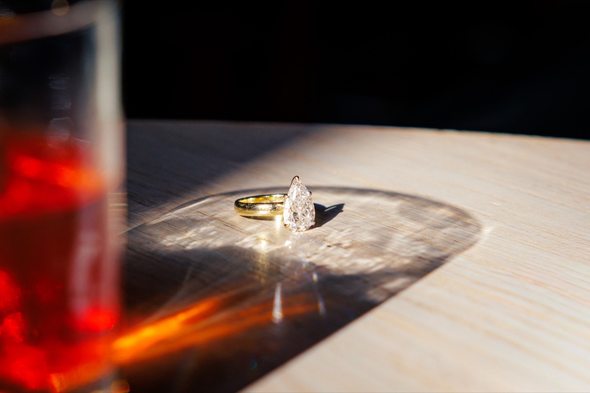 A pear-cut diamond engagement ring on a sunlit table beside a glass of red