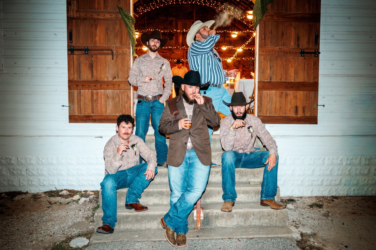 Groomsmen in cowboy hats light cigars on the steps of a barn