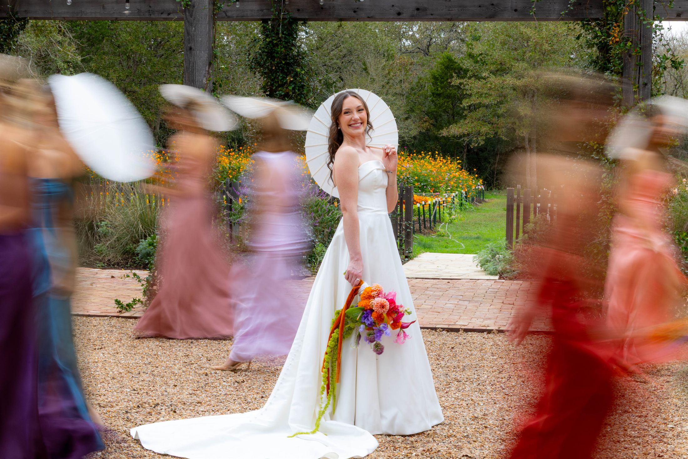 A bride smiles beneath a white parasol as her bridesmaids blur past in color