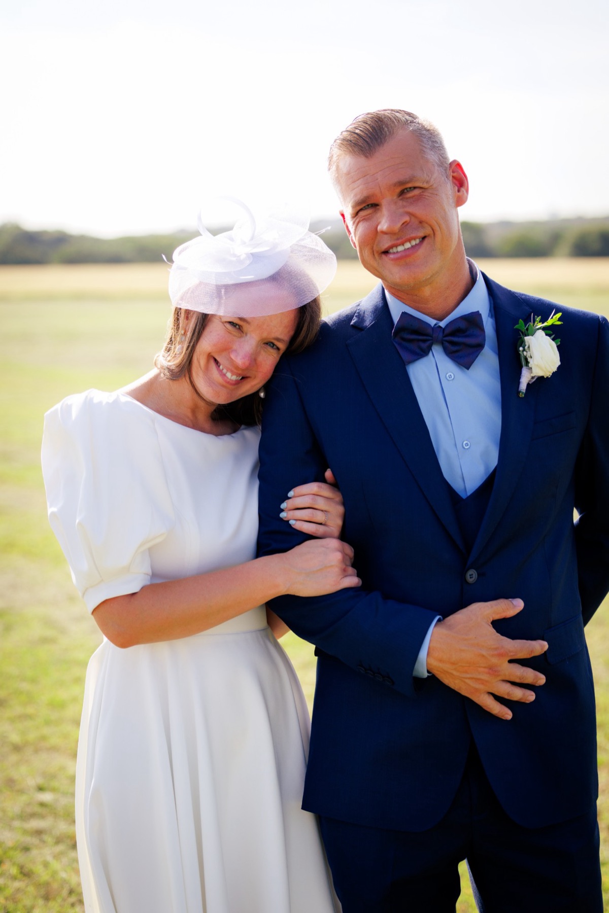 A bride in a white fascinator leans her head on her groom's shoulder in an open field